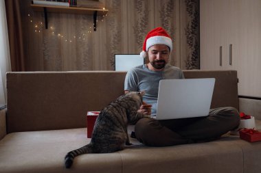 Noel zamanı evdeki kanepede oturan gri kedili hippi bir adam. Sanal Noel partisi. Çevrimiçi takım toplantı videosu evden arıyor. Noel Baba şapkalı adam