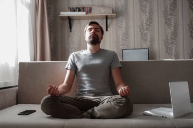 Milenyum hippi erkeği evdeki kanepede rahatça dinlenemiyor. Erkekler kanepede dizüstü bilgisayarla ve telefonla meditasyon yaparlar.