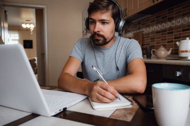 Odaklanmış genç adam internetten kulaklık takıyor laptopta webinar podcast izliyor eğitim konferansını dinliyor notlar alıyor çalışma masasında oturuyor, konsept öğreniyor.