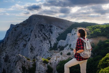 Sırt çantalı ve sırıklı formda bir kadın yürüyüşçü kayalık bir dağ sırtında dikilip sağlıklı bir doğa yaşam tarzı konseptiyle tepelere bakıyor.