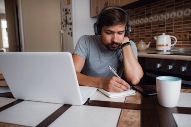 Odaklanmış genç adam internetten kulaklık takıyor laptopta webinar podcast izliyor eğitim konferansını dinliyor notlar alıyor çalışma masasında oturuyor, konsept öğreniyor.