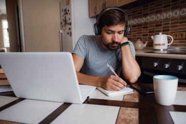 Odaklanmış genç adam internetten kulaklık takıyor laptopta webinar podcast izliyor eğitim konferansını dinliyor notlar alıyor çalışma masasında oturuyor, konsept öğreniyor.