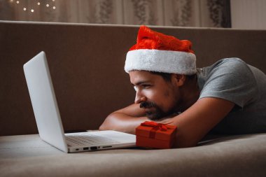 Noel zamanı evdeki kanepede oturan kırmızı bardaklı hippi bir adam. Sanal Noel partisi. Çevrimiçi takım toplantı video konferansı evden arıyor..