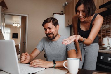 Genç bir çift mutfakta dizüstü bilgisayar ve akıllı telefon kullanıyor, evde çalışan gülümseyen bir adam, alet ya da cihaz bağımlılığı olan aile sabahı yaşam tarzı. okuma haberleri