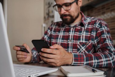 Akıllı telefonla ödeme yapmak için kredi kartını kullanan yakışıklı bir adam. İnternetten alışveriş, bankacılık sistemi. Ekose gömlekli bir adam mutfağında oturuyor.