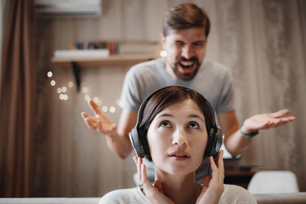 angry husband shouting at wife sitting on sofa and covering ears with headphones. the woman ignores the man