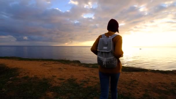 身穿黄色毛衣 头戴红色帽子的女人 从海角俯瞰着美丽多彩的落日 胜利者的概念 徒步旅行者探索自然 — 图库视频影像