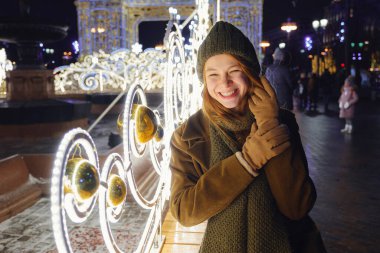 Sokakta poz veren gülümseyen genç bir kadın. Arka planda Noel festivali var. Şık bir kışlık palto, örgü şapka ve eşarp giyen bir manken. Büyük şehirde mutlu hissediyorum. Moskova 'da kış tatili