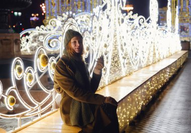 Sokakta poz veren gülümseyen genç bir kadın. Arka planda Noel festivali var. Şık bir kışlık palto, örgü şapka ve eşarp giyen bir manken. Büyük şehirde mutlu hissediyorum. Moskova 'da kış tatili