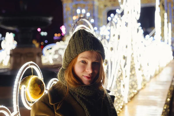 路上でポーズしている若い笑顔の女性 背景にあるお祭りのクリスマスフェア スタイリッシュな冬のコート ニットキャップ スカーフを身に着けているモデル 大都市で幸せな気分 冬の休暇を過ごすモスクワ — ストック写真