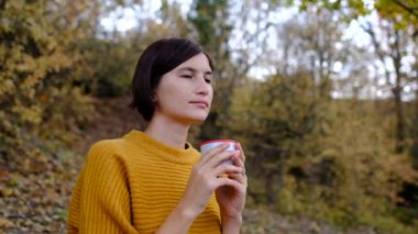 Sıcak bir içkiyle genç ve güzel bir kadın sonbahar ormanlarında tatil yapıyor..