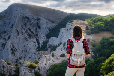 Sırt çantalı ve sırıklı formda bir kadın yürüyüşçü kayalık bir dağ sırtında dikilip sağlıklı bir doğa yaşam tarzı konseptiyle tepelere bakıyor.