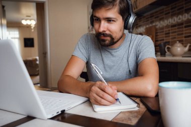 Odaklanmış genç adam internetten kulaklık takıyor laptopta webinar podcast izliyor eğitim konferansını dinliyor notlar alıyor çalışma masasında oturuyor, konsept öğreniyor.