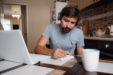 Odaklanmış genç adam internetten kulaklık takıyor laptopta webinar podcast izliyor eğitim konferansını dinliyor notlar alıyor çalışma masasında oturuyor, konsept öğreniyor.
