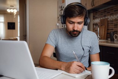 Odaklanmış genç adam internetten kulaklık takıyor laptopta webinar podcast izliyor eğitim konferansını dinliyor notlar alıyor çalışma masasında oturuyor, konsept öğreniyor.