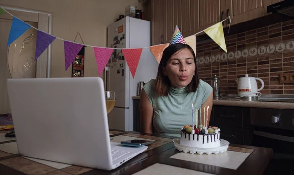 Woman blowing out the candle on the birthday cake and making video call. Lady celebrating birthday online in quarantine time. Authentic decorated home workplace. Coronavirus outbreak 2020.
