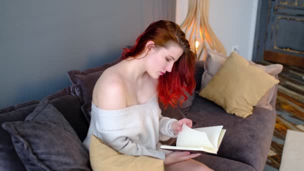 Young woman siting on a sofa at home. reading a book thoughtfully and dreamily. — Stock Video