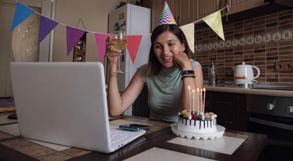 Woman celebrating her birthday through video call virtual party with friends. Lits and blows out candle. Authentic decorated home workplace. Coronavirus outbreak 2021.