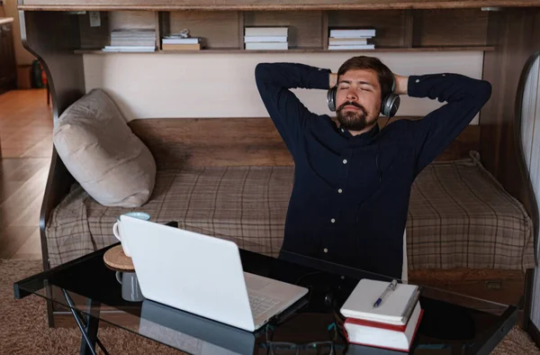 Satisfied businessman wearing headphones enjoying music with closed eyes, relaxing during break, sitting at work desk, smiling young man wearing glasses listening to favorite song