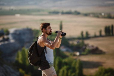Tepedeki dürbüne bakan adam. Sırt çantalı bir adam. Vadi manzarasında yürüyüş yapan genç beyaz bir adam.