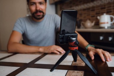 Genç bir adam, ofisteki akıllı telefondan video görüşmesi yapıyor. Sanal ev partisi. Kendine güvenen genç vlogger etkileyicisi evde sosyal medya video blogu konuşması çekiyor