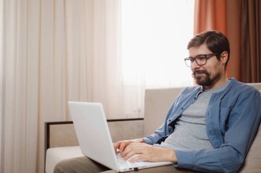 Evde dizüstü bilgisayar kullanan yakışıklı bir genç. Öğrenci erkekler odasında. Çevrimiçi alışveriş, ev işi, serbest çalışma, çevrimiçi öğrenme, öğrenim kavramı. Uzaklık eğitimi