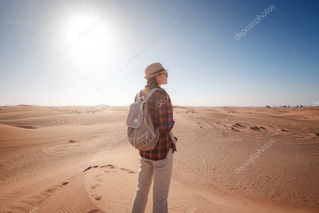 attractive asian young woman in plaid shirt in desert, treveling in UAE on safari, wearing hat and backpack, exploring nature of sandy beauty