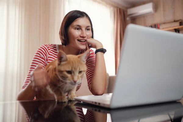 Happy young woman in a white and red tshirt works at home with a laptop and a ginger cat, remote work and education, staying home during a coronavirus
