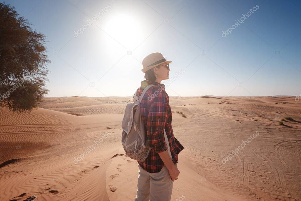 attractive asian young woman in plaid shirt in desert, treveling in UAE on safari, wearing hat and backpack, exploring nature of sandy beauty