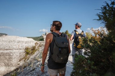 İki adam kayalık yamaçlarda yürüyüş çubuklarını kullanarak sırt çantalarını taşıyorlar. Dağlar Vadisi, insanların yanı sıra manzaralı. Doğaya yaz seyahati. Arka planda yürüyüşçü.
