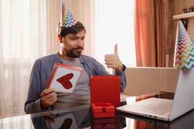 Mutlu genç adam masada oturup arkadaşlarıyla görüntülü konuşma yapıyor. Bir erkek hediyesini açar, onunla çok mutlu olur.