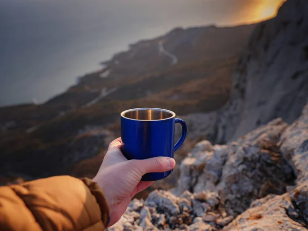 Closeup photo of cup with tea in traveler\'s hand over out of focus mountains view. A tourist man drink a hot tea from cup and enjoys the scenery in the mountains. Trekking concept