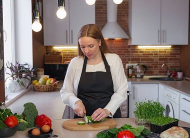 Sağlıklı yemek pişirirken mutfakta duran mutlu, genç bir sarışın bayan. Sağlıklı Yaşam Tarzı Konsepti. Evde yemek pişirmek. Yemek yapmak için sebze doğrama