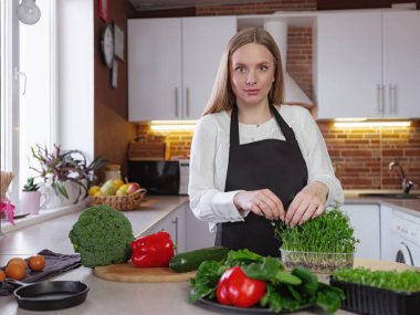 Sağlıklı yemek pişirirken mutfakta duran mutlu, genç bir sarışın bayan. Sağlıklı Yaşam Tarzı Konsepti. Evde yemek pişirmek. Sağlıklı bağırsak mikroflorasının garantisi olarak kabın içindeki mikroyeşiller.
