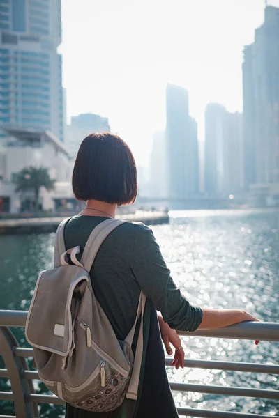 Happy young female traveler in the big city of Dubai, famous place Dubai marina. Luxury and comfortable tourism season in United Arab Emirates. Back or rear view of young woman in dress and backpack