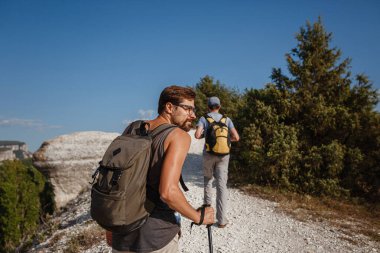 İki adam kayalık yamaçlarda yürüyüş çubuklarını kullanarak sırt çantalarını taşıyorlar. Dağlar Vadisi, insanların yanı sıra manzaralı. Doğaya yaz seyahati. Arka planda yürüyüşçü.
