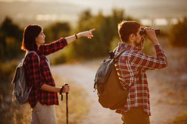 Yürüyüş gezilerinde eğlenen genç çiftin portresi. Kafkasyalı ve Asyalı yürüyüşçü çift yaz tatilinde eğleniyorlar. Doğayı dürbünle izliyorlar..