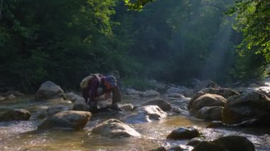 Erkek gezgin gün batımında ormanda nehrin kıyısında doğanın güzelliğinin tadını çıkarıyor. Sinema ve ilham verici seyahat blogcusu canlı motivasyon macerası. Adam nehirden su içiyor.