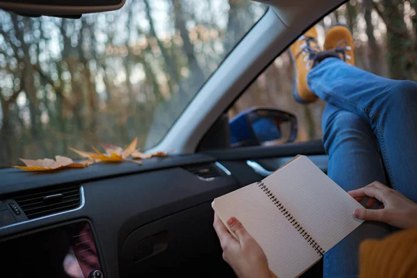 Modern hipster woman resting in a car and reading. . Feet outside the window at sunset forest. The concept of freedom of movement. An autumn weekend in nature.