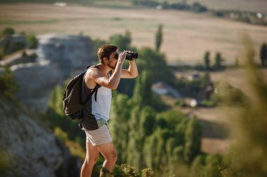 Tepedeki dürbüne bakan adam. Sırt çantalı bir adam. Vadi manzarasında yürüyüş yapan genç beyaz bir adam.
