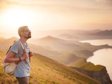 Sırt çantalı genç sporcu kayanın tepesinde duruyor ve yazın günbatımında deniz kıyılarına ve dağlara bakıyor. Açık havada seyahat etmenin yaşam tarzı konsepti.