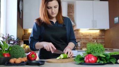 Ev yemeği ve evde yemek pişirme deneyimi konsepti. Siyah önlüklü kızıl saçlı bir kadın Chukini 'yi Shakuka için mutfağında kesiyor.