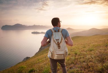 Sırt çantalı genç sporcu kayanın tepesinde duruyor ve yazın günbatımında deniz kıyılarına ve dağlara bakıyor. Açık havada seyahat etmenin yaşam tarzı konsepti.