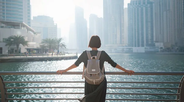 Happy young female traveler in the big city of Dubai, famous place Dubai marina. Luxury and comfortable tourism season in United Arab Emirates. Back or rear view of young woman in dress and backpack