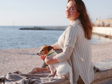 Jack Russell Terrier 'ı olan genç bir kadın. Sevimli hayvan. Bir kadın ve sadık evcil hayvanı deniz kenarında günbatımının tadını çıkarırlar.