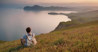 Sırt çantalı bir gezgin bir dağın tepesinde oturur ve kıyı manzarasının tadını çıkarır. Dağ ve kıyı seyahati, özgürlük ve aktif bir yaşam tarzı