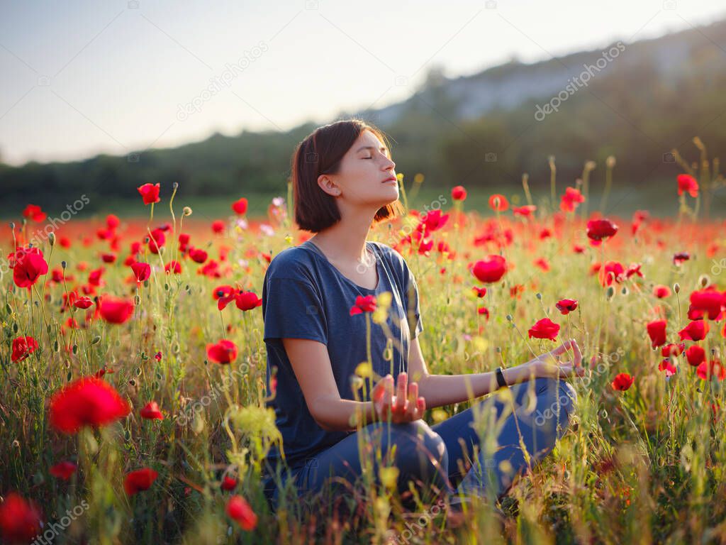 A beautiful woman meditates on a poppy field at sunset. Wellness well-being happiness concept.