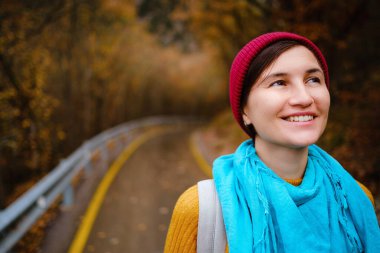 Kırmızı şapkalı güzel bayan hippi, sarı kazak ve mavi atkı sonbahar doğasında yürüyor. Hava serin. Gezgin fotoğraf serisi.