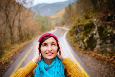 Kırmızı şapkalı güzel bayan hippi, sarı kazak ve mavi atkı sonbahar doğasında yürüyor. Hava serin. Gezgin fotoğraf serisi.