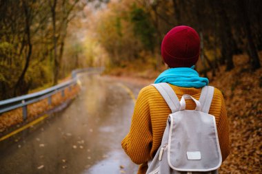 Kırmızı şapkalı güzel bayan hippi, sarı kazak ve mavi atkı sonbahar doğasında yürüyor. Hava serin. Gezgin fotoğraf serisi.